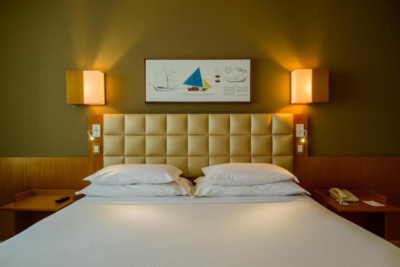 Hotel Luzeiros Sao Luis Extérieur photo The photo shows a modern hotel room featuring a large bed with a tufted beige headboard. There are two white pillows on the bed and a smooth white bedspread. On the wall above the bed, there's a framed artwork with a colorful sailboat and nautical de