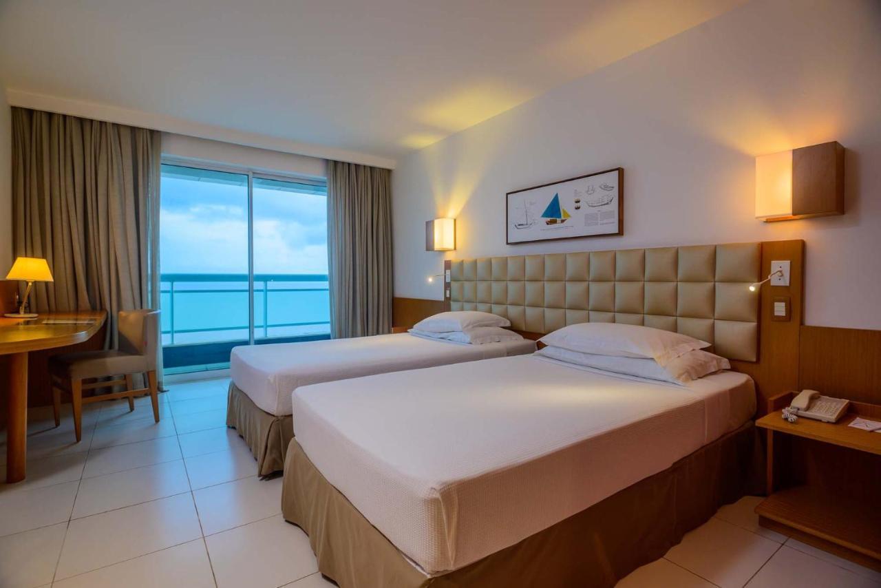 Hotel Luzeiros Sao Luis Extérieur photo The photo shows a hotel room with two single beds. The beds are neatly made, featuring light-colored bedding. Behind the beds is a beige upholstered headboard. There are two lamps mounted on the wall above the headboard and a piece of artwork featuri