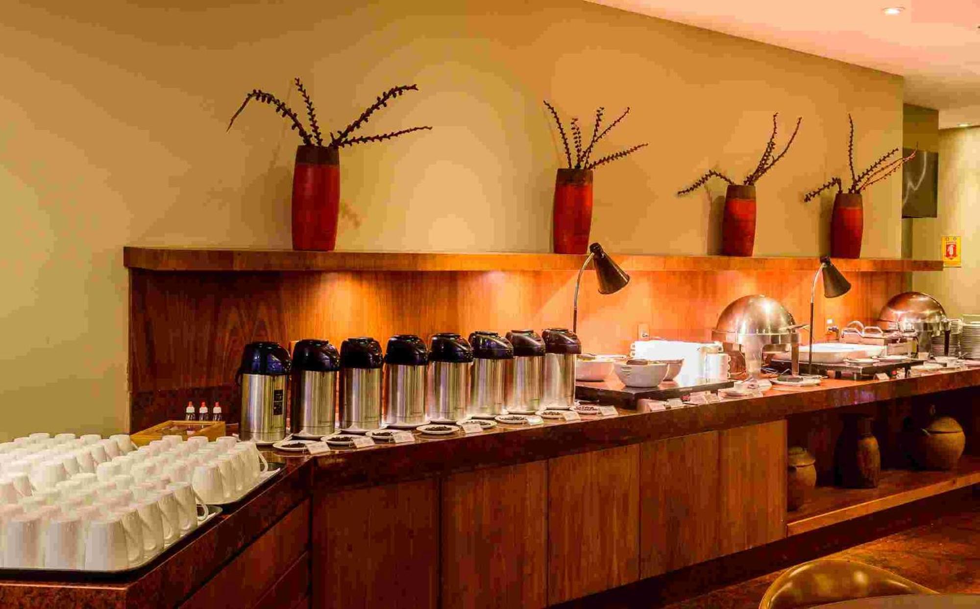 Hotel Luzeiros Sao Luis Extérieur photo The photo shows a buffet setup in a dining area. There is a long wooden counter that features several items:

1. **Beverages**: Multiple stainless steel coffee or tea containers are lined up, suggesting a beverage station.
2. **Dishware**: There are 