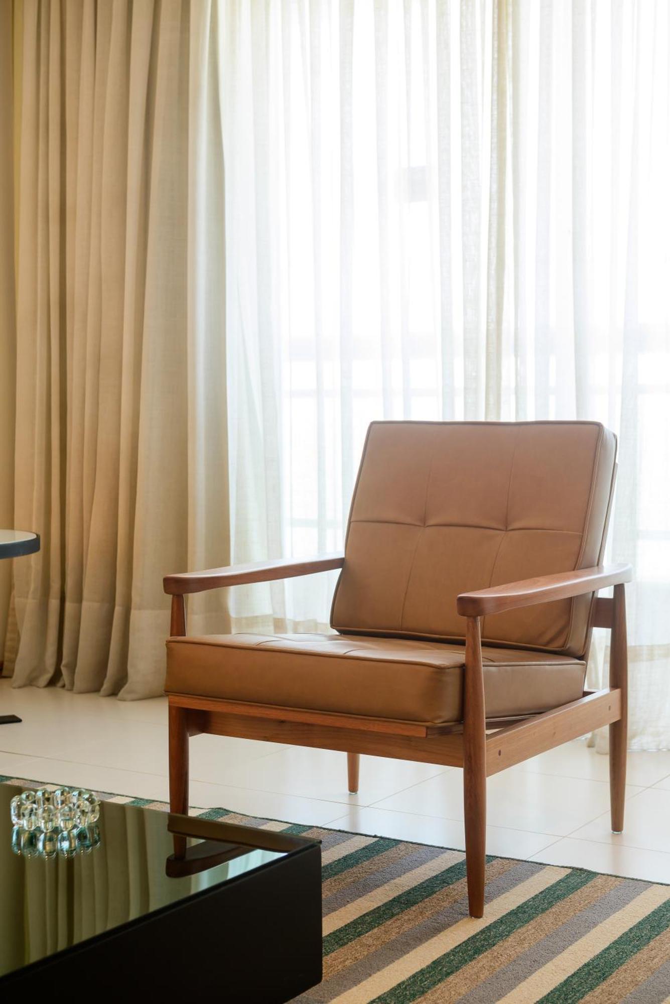 Hotel Luzeiros Sao Luis Extérieur photo The photo shows a stylish, minimalist armchair with a simple wooden frame and a tan leather cushion. It is positioned near large curtains that allow natural light to enter the room, creating a warm and inviting atmosphere. The overall setting appears