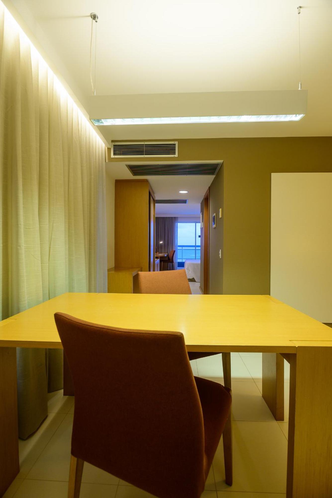 Hotel Luzeiros Sao Luis Extérieur photo The photo shows the interior of a room, likely a hotel or apartment. In the foreground, there is a wooden table with a chair. The walls are painted in a warm brown color, and there are large curtains that partially cover a window. Beyond the table, t
