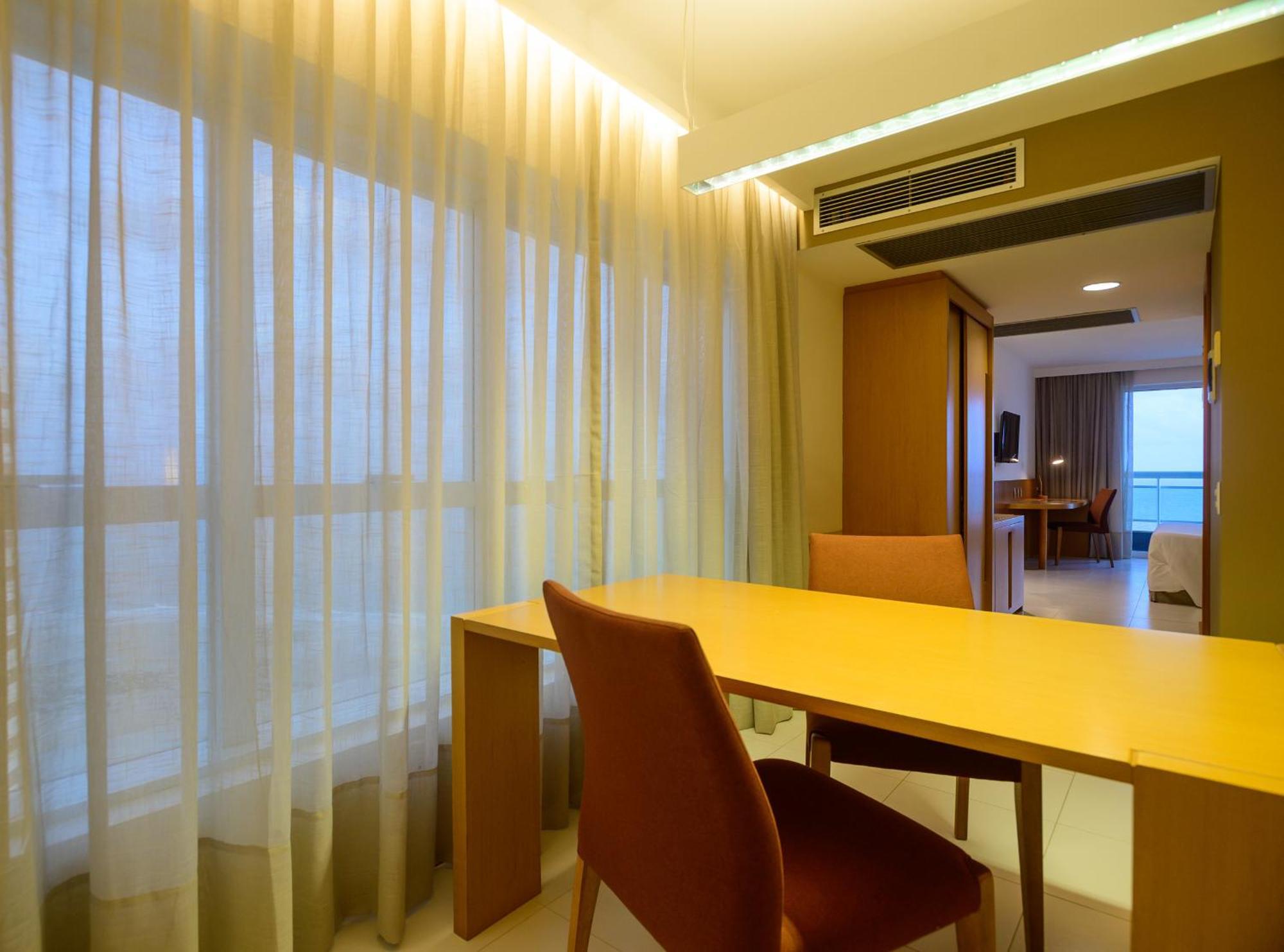 Hotel Luzeiros Sao Luis Extérieur photo The photo shows a modern, well-lit interior space, likely a hotel room or a living area. There is a wooden table with two chairs positioned in the foreground. The table is simple and functional, while the chairs have a soft, cushioned seat and slight